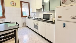 Una cocina con armarios blancos, microondas, refrigerador, pileta y ventana con persianas.
