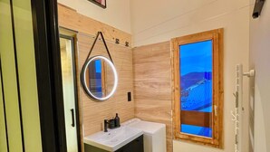Une salle de bain moderne avec un meuble-lavabo en bois, un miroir rond, une fenêtre offrant une belle vue et un plafond avec des poutres apparentes.