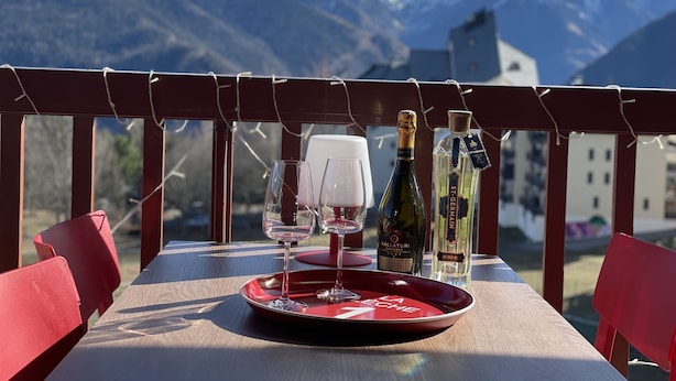 Une table avec un plateau rouge, deux verres à vin, une bouteille de Ballaturis et une bouteille de vin St. Germain, le tout disposé devant un paysage de montagne.