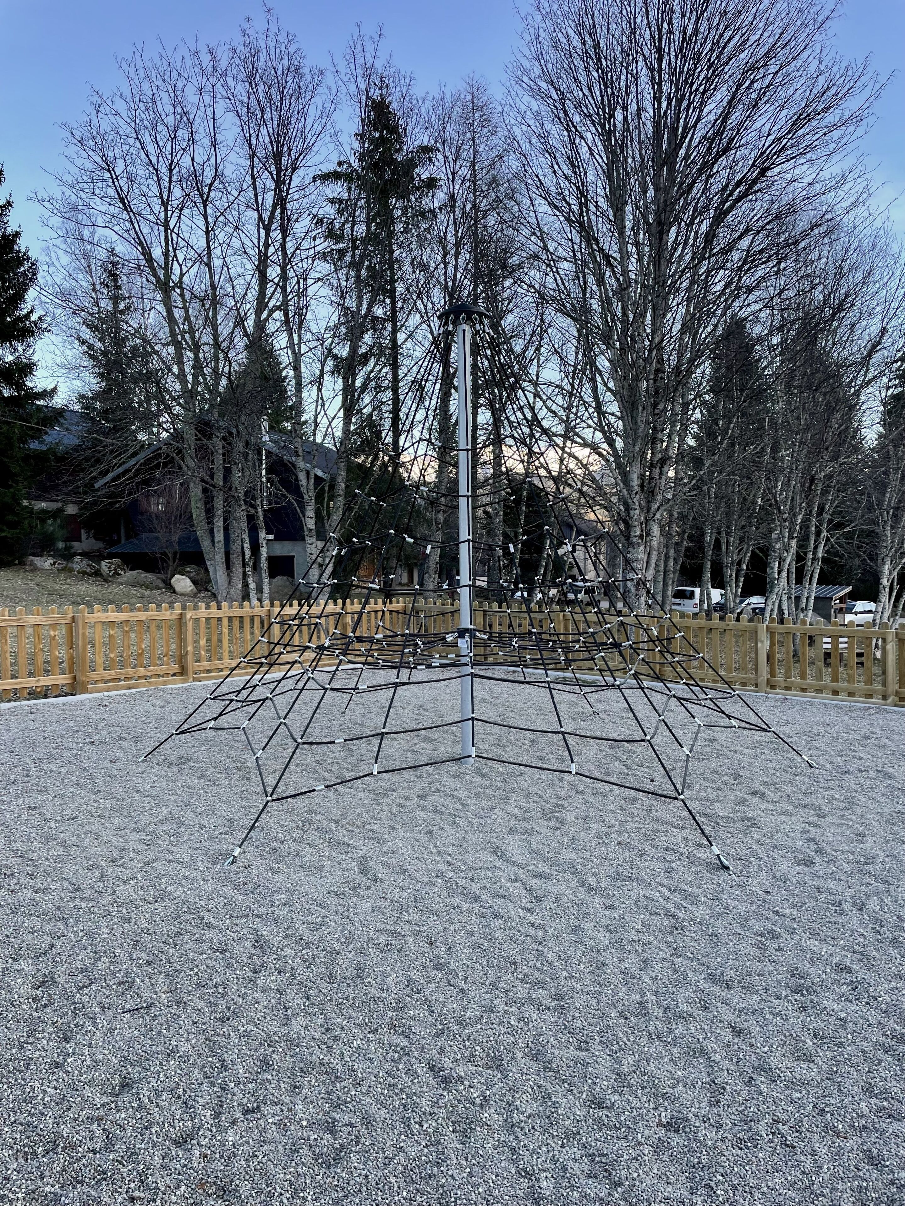Un parque infantil con una estructura para escalar, rodeado de árboles y una cerca de madera.
