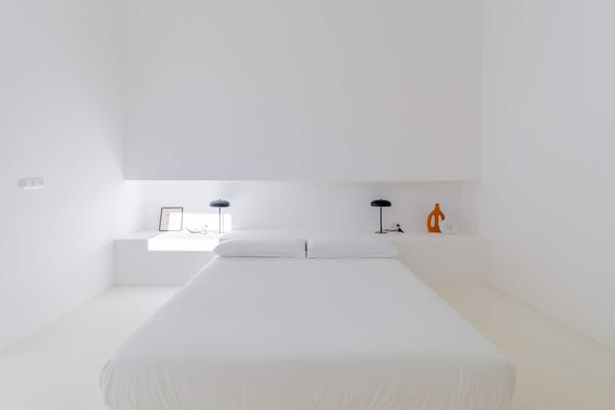 Een minimalistische slaapkamer met een groot bed, een nachtkastje, een lamp en een klein oranje sculptuur.