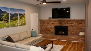 Una sala de estar con chimenea de ladrillo, un sofá, un televisor y tres pinturas de paisajes en la pared.