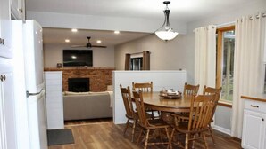Un comedor con mesa y sillas de madera, un televisor sobre la chimenea y un ventanal con cortinas.