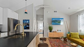 Eine moderne Küche mit schwarzem Arbeitsplatz, einem Edelstahlkühlschrank und einem goldenen Wasserhahn. Es gibt einen Essbereich mit Holzboden, einem farbenfrohen Teppich und einem gelben Sofa.