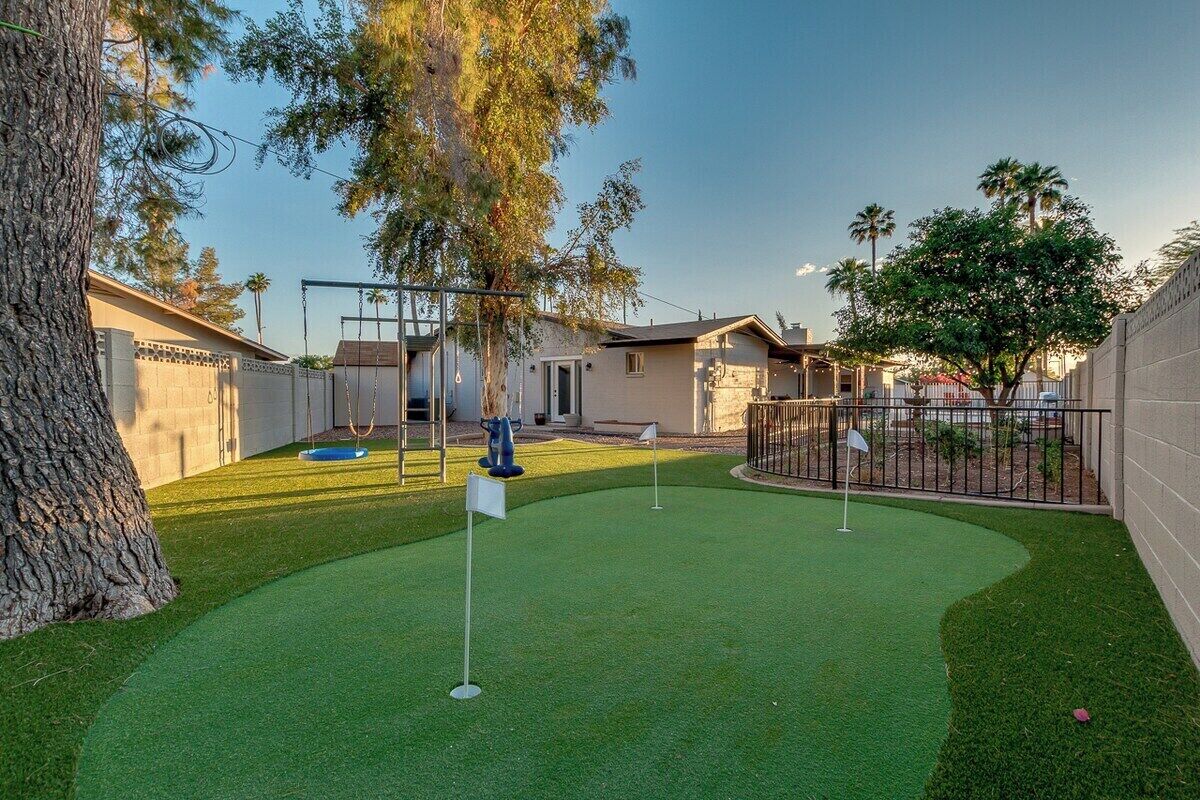 A backyard with artificial turf, a swing set, and a fenced area with trees and a house in the background.