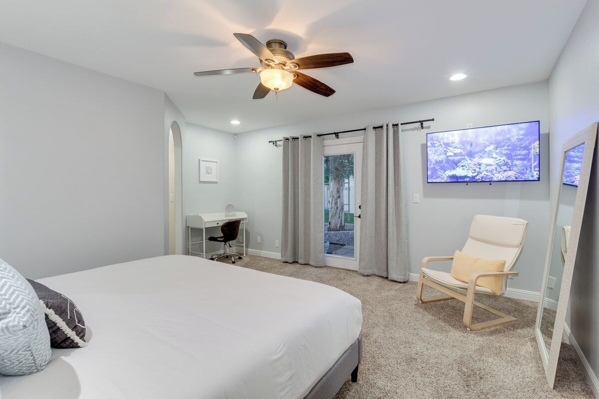 Une chambre avec un lit, un bureau, une chaise, un ventilateur de plafond, une télévision et une porte coulissante donnant sur un balcon.