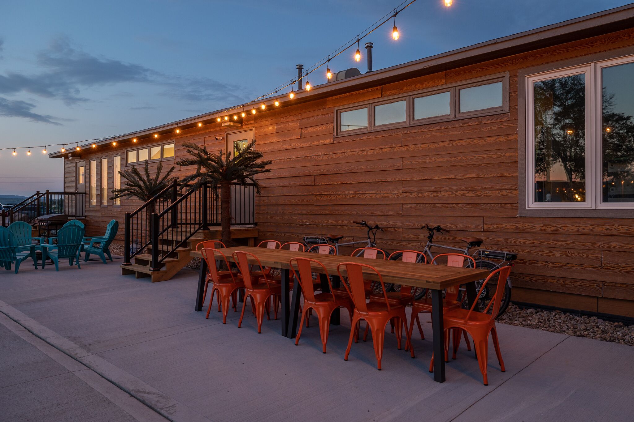 A wooden building with a dining area featuring a long table and chairs, a staircase, and string lights.