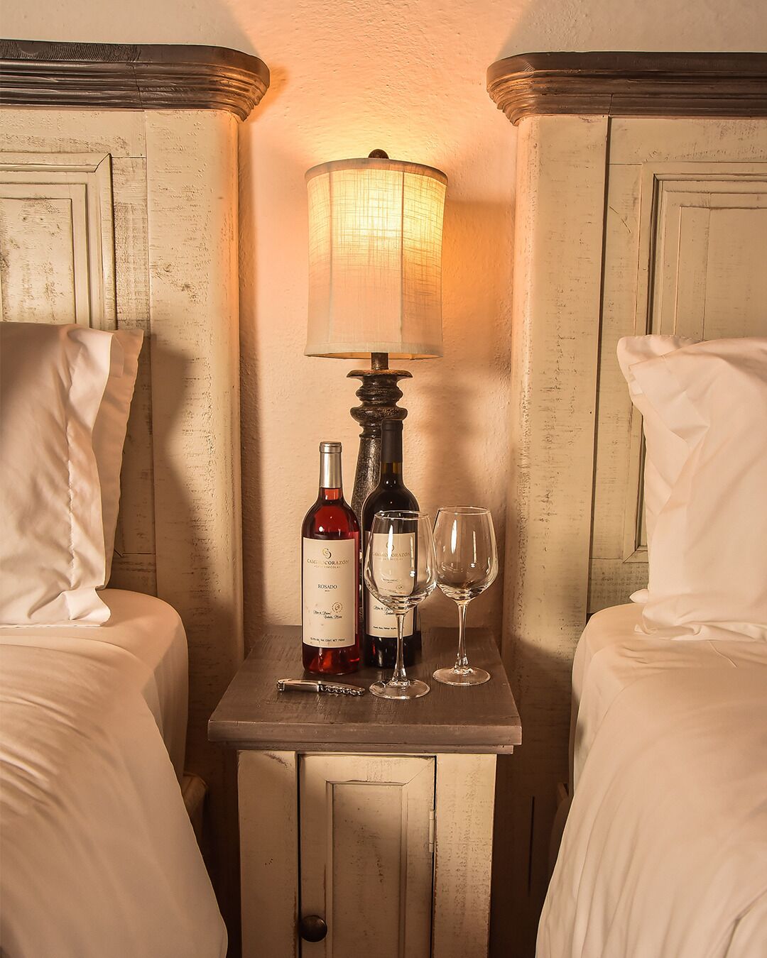 Quarto de hotel com duas camas, uma mesa de cabeceira com abajur, duas garrafas de vinho e dois copos de vinho.