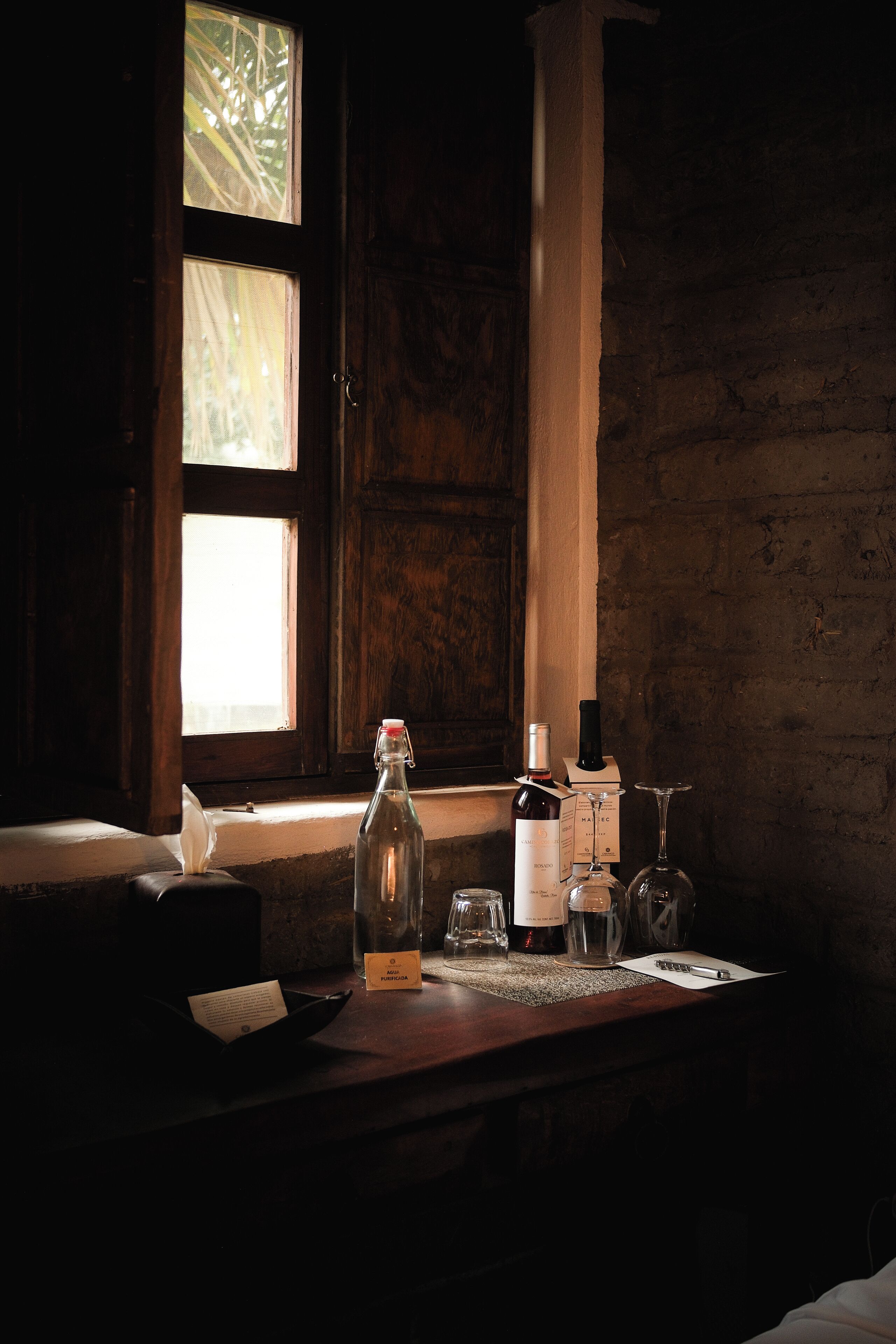 A dimly lit room with a wooden table, a bottle, a wine glass, and a window with sunlight.