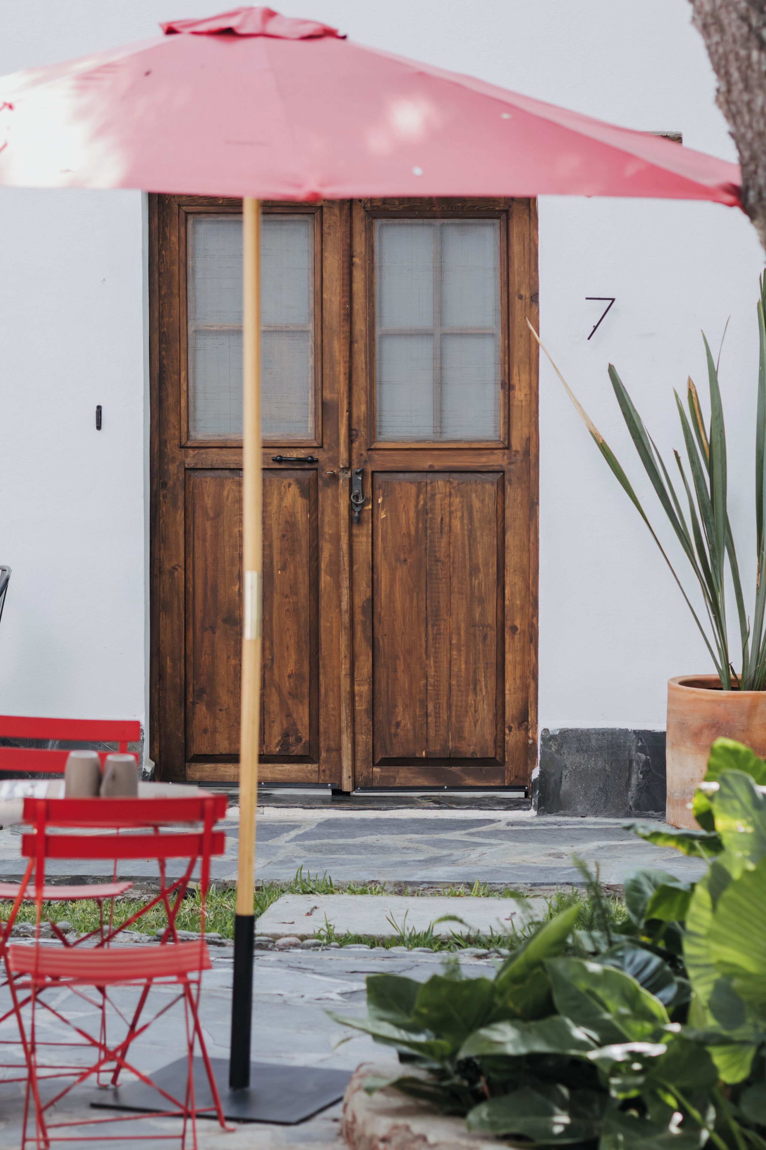A wooden double door with a number 7, a red umbrella, and a red chair outside.