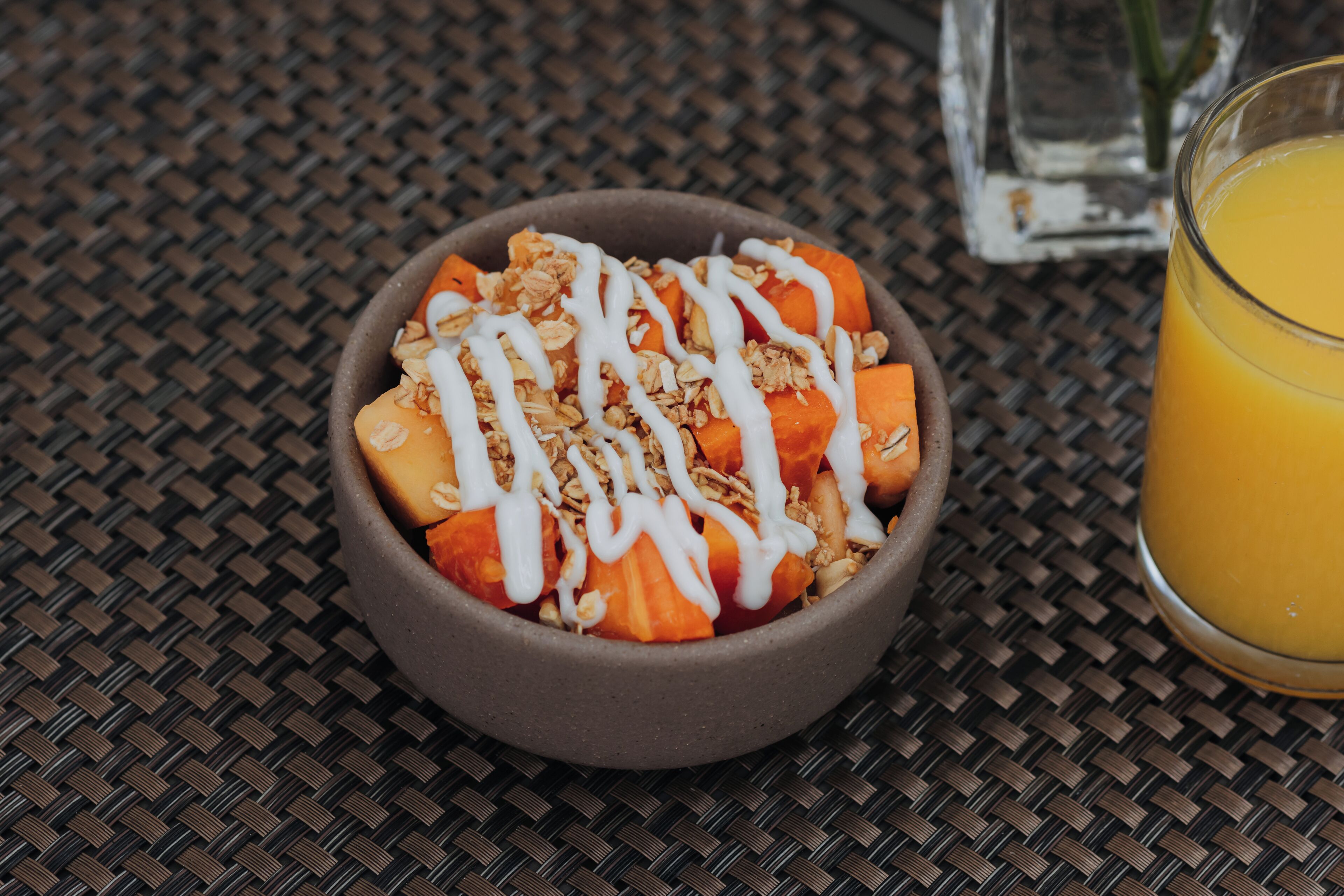 A bowl of fruit salad with granola and white syrup, accompanied by a glass of orange juice on a woven table.