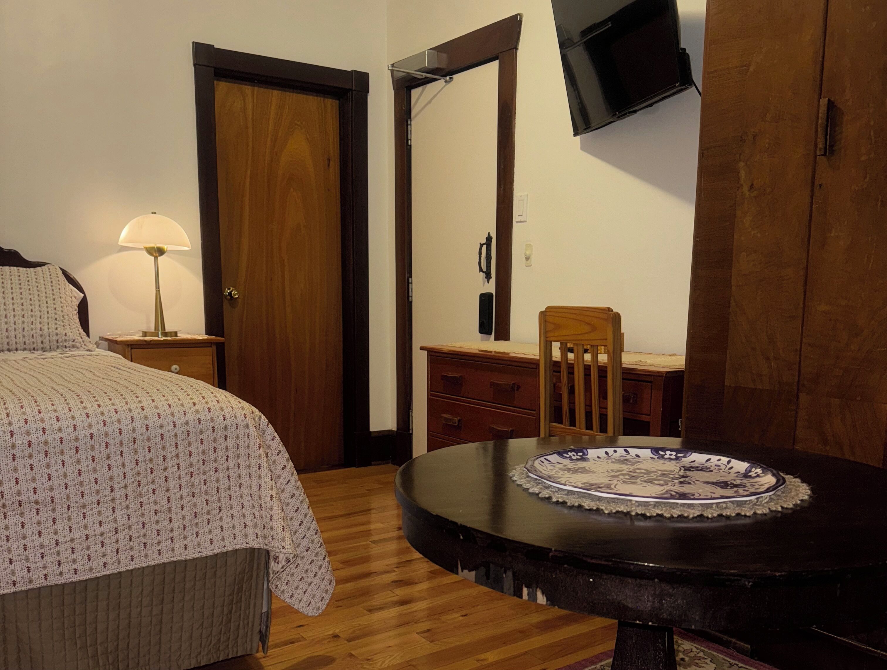 A bedroom with a bed, bedside lamp, dresser, chair, and a round table with a plate.