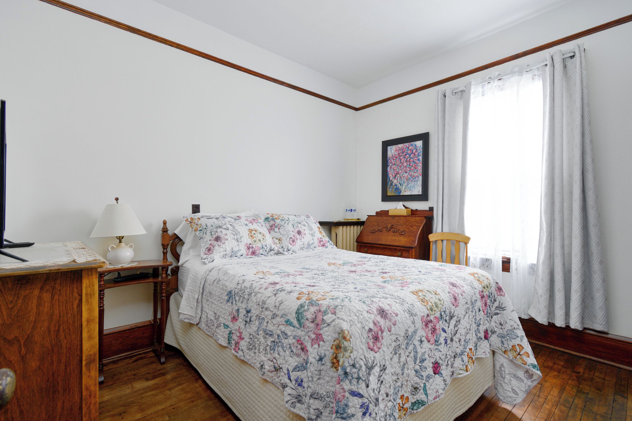 Un dormitorio con cama, escritorio, lámpara, silla, radiador y ventana con cortinas.