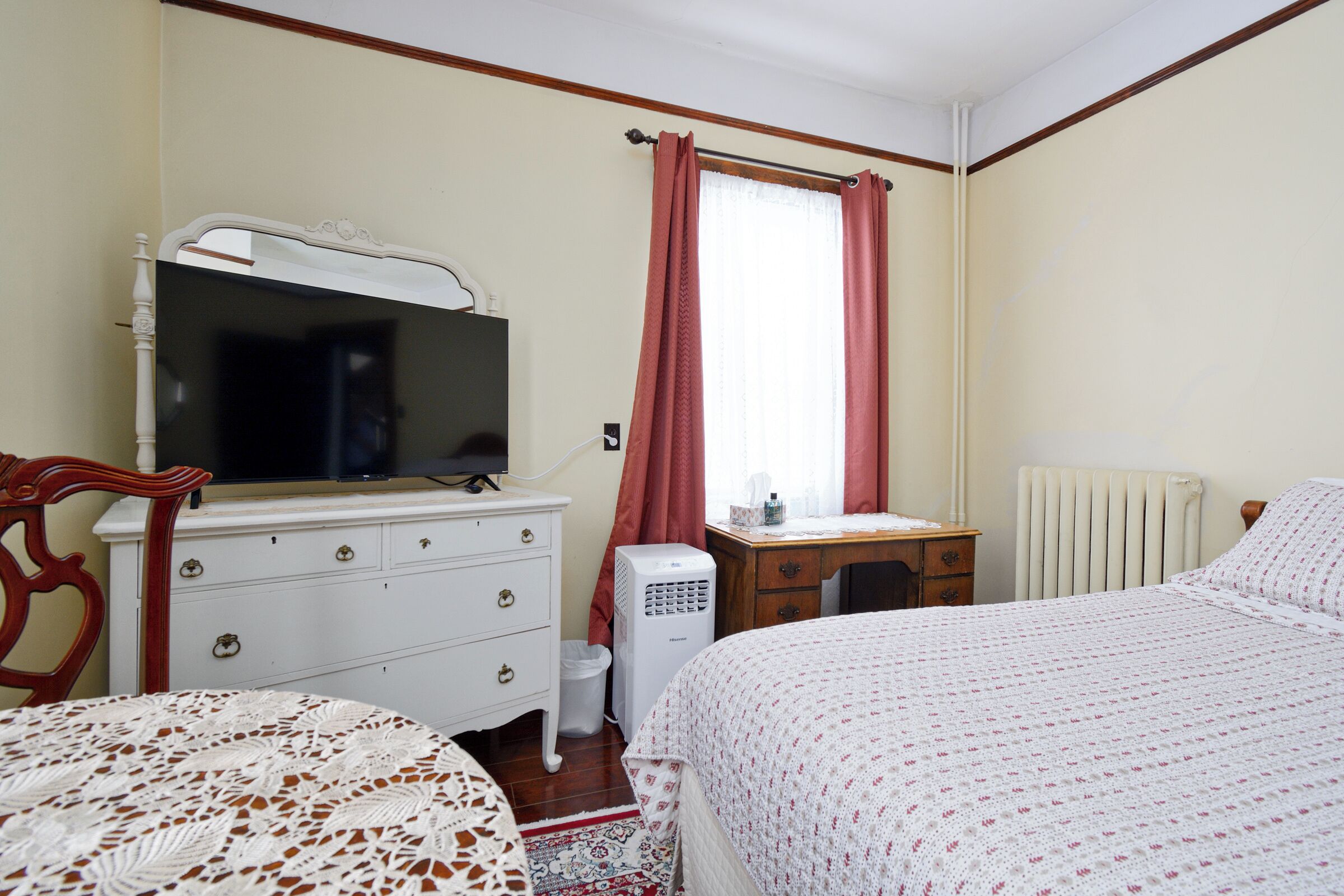 A bedroom with a bed, a dresser, a desk, a chair, a television, and a radiator.