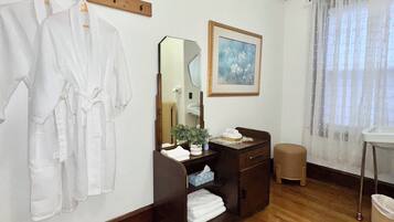 A hotel room with a wooden floor, a wooden bench, a mirror, a sink, a framed picture, and a window with sheer curtains.