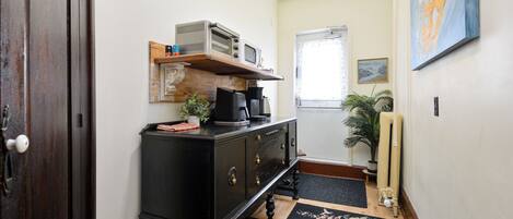 Un pasillo estrecho con una puerta de madera, un mueble negro con un microondas encima, un estante pequeño con objetos decorativos, una planta en maceta y un radiador.