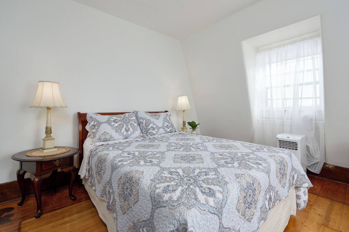 A bedroom with a large bed, two bedside tables with lamps, a window with sheer curtains, and a radiator.