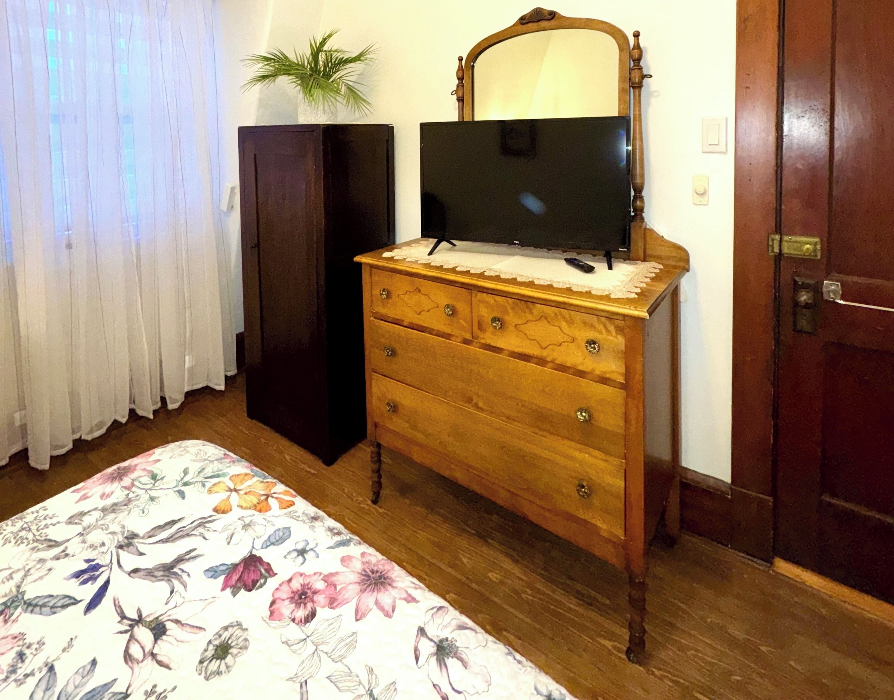 Ein Schlafzimmer mit einem Holzkoffer, einem Fernseher darauf, einer Topfpflanze, einer blumigen Tagesdecke und einer Holztür.