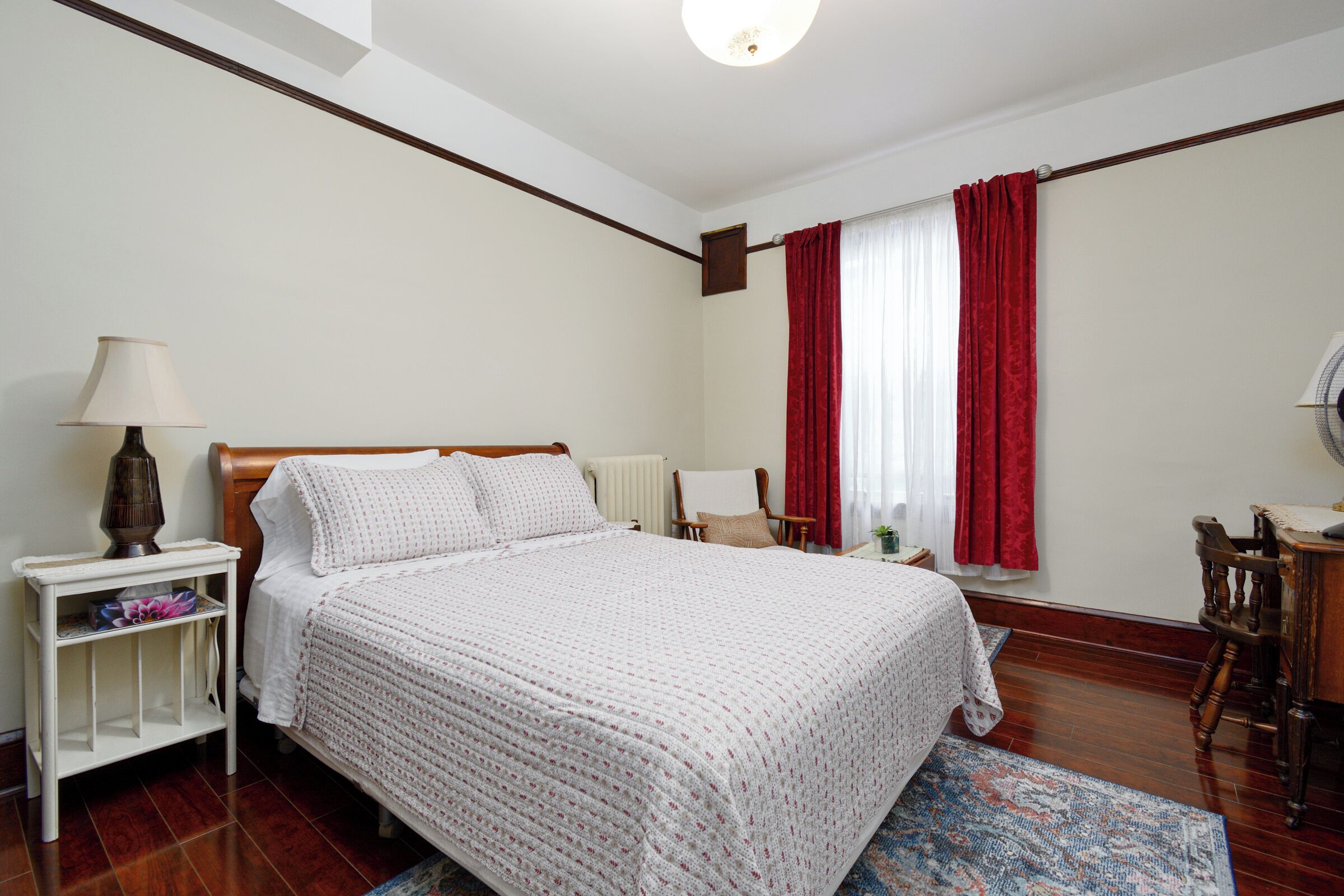 A bedroom with a wooden bed, white bedding, a nightstand with a lamp, a chair, and a window with red curtains.