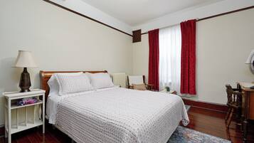 A bedroom with a wooden bed, white bedding, a nightstand with a lamp, a chair, and a window with red curtains.