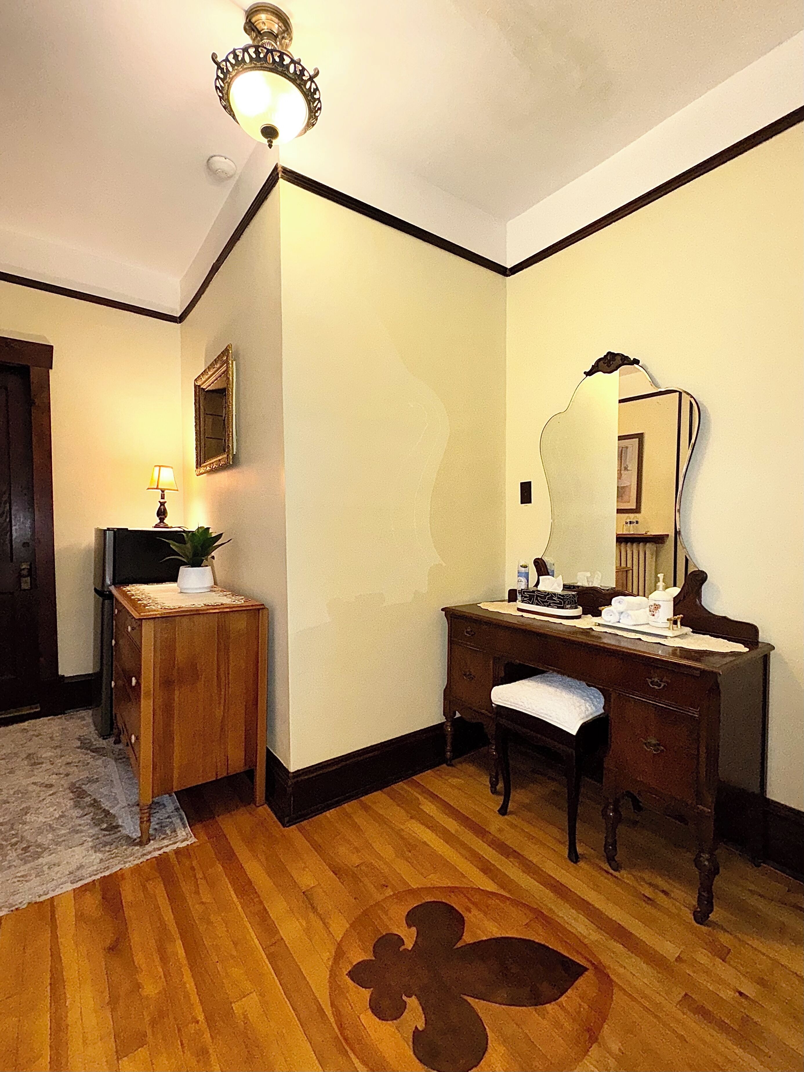 A room with a wooden dresser, a vanity with a mirror, a small table with a lamp, and a floral rug.
