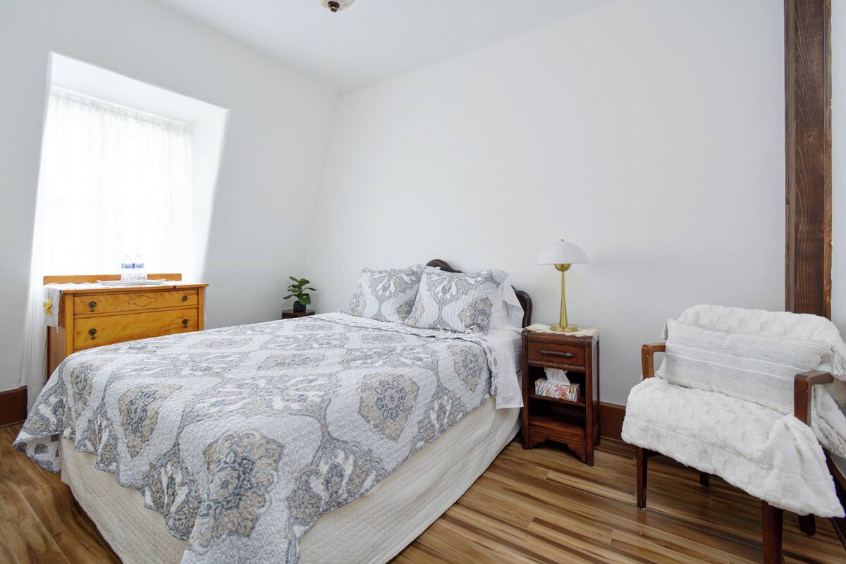 A bedroom with a large bed, wooden nightstand, bedside table, and a chair.
