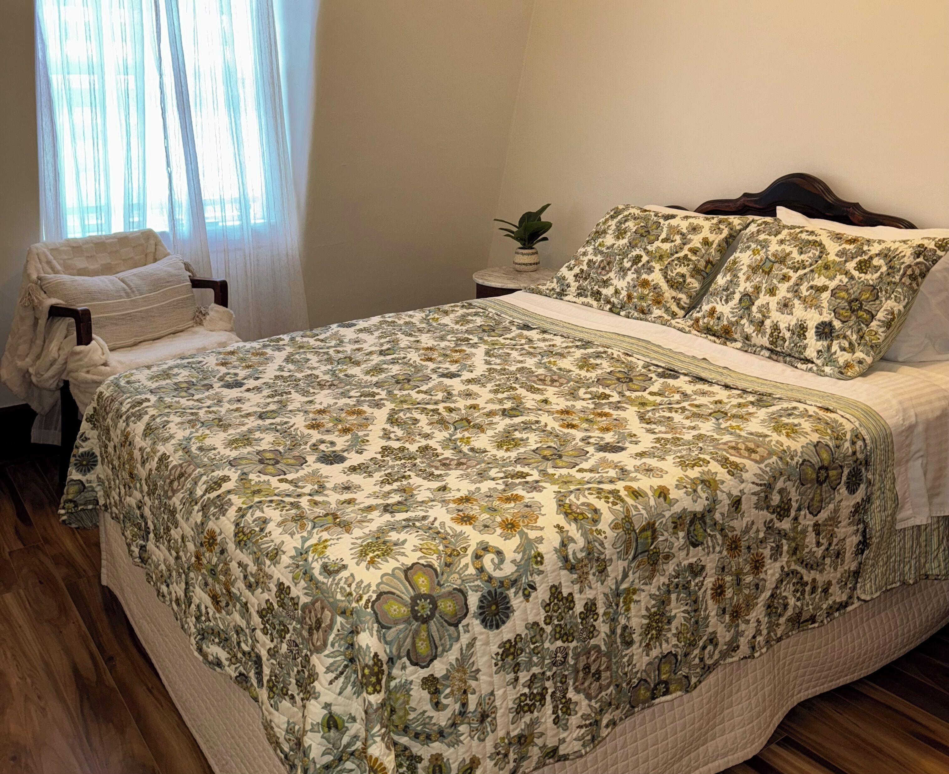 Une chambre à coucher avec une couvre-lit à motifs floraux, une table de chevet ornée d’une plante et une fenêtre avec des rideaux légers.