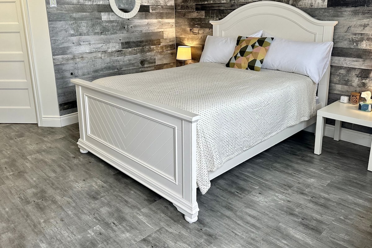 Un dormitorio con una cama blanca, una mesita de noche, una lámpara y una pared de madera.