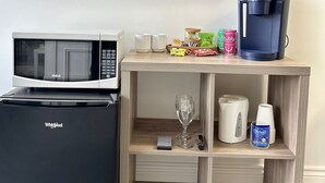 Uma cozinha compacta com micro-ondas, geladeira, cafeteira e uma pequena prateleira de madeira com xícaras, um copo e uma garrafa.