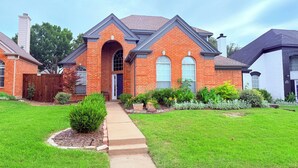 A two-story brick house with a front entrance, a covered porch, and a well-maintained lawn. - Frisco Resort Style Home with Spa (Frisco)