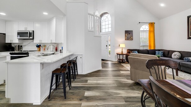 A modern kitchen with white cabinetry, a central island with bar stools, a dining area with a wooden table, and a living room with a sofa and a lamp. - Frisco Resort Style Home with Spa (Frisco)
