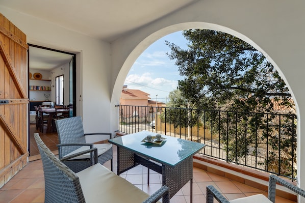 A balcony with wicker furniture, a glass table, and a view of a tree and buildings.