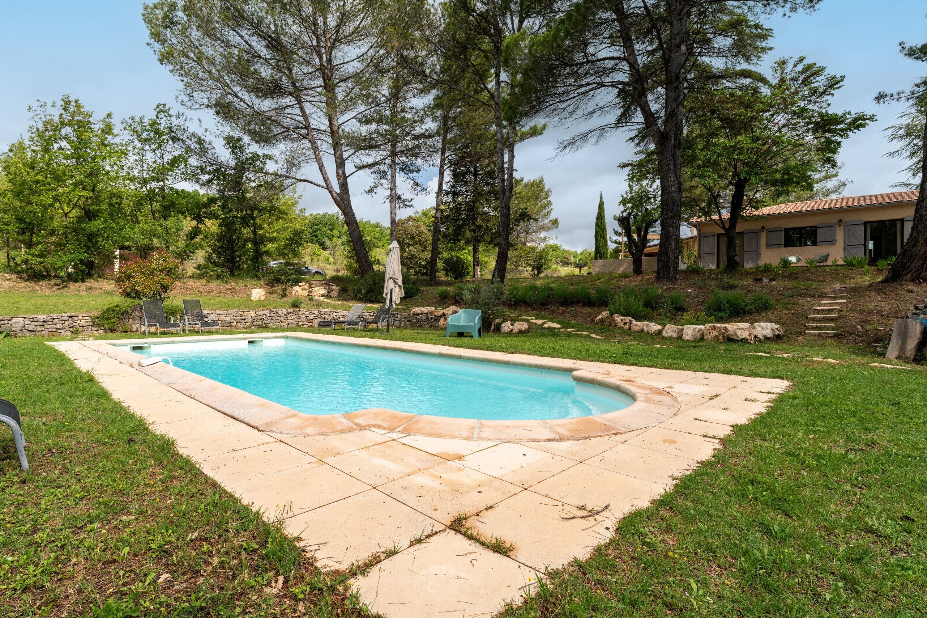 A swimming pool with a stone deck, surrounded by grass and trees, with a stone wall and a building in the background.