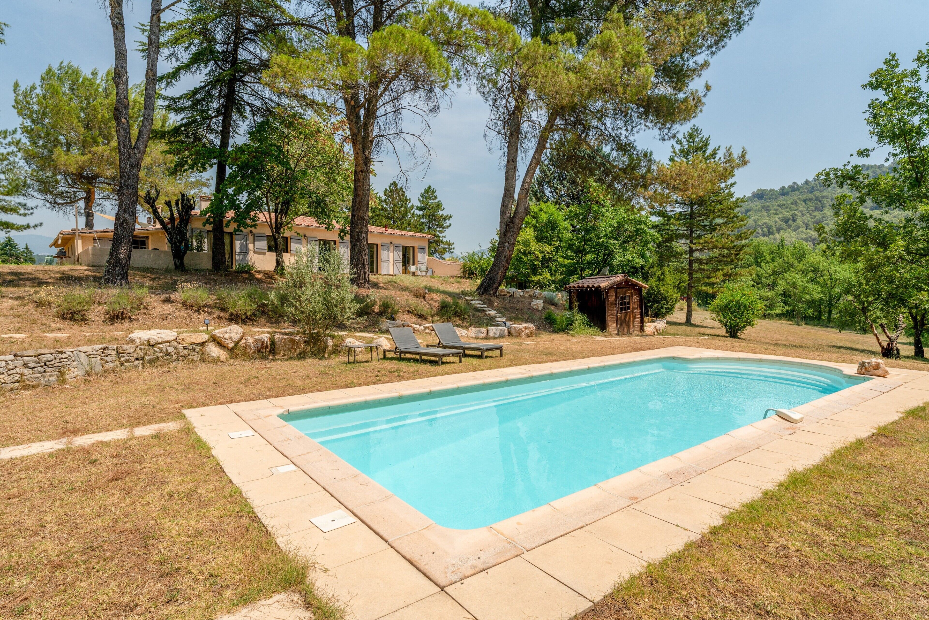 A swimming pool with lounge chairs, surrounded by trees and a stone wall.
