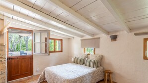 A bedroom with a bed, a wooden stool, a window with a view of trees, and a stone wall.