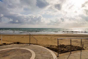 A beach with a curved stone pathway leading to the water, metal railings, and a cloudy sky.