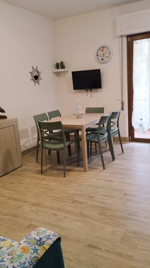 A dining area with a wooden table and four chairs, a television mounted on the wall, and a small shelf with decorative items.