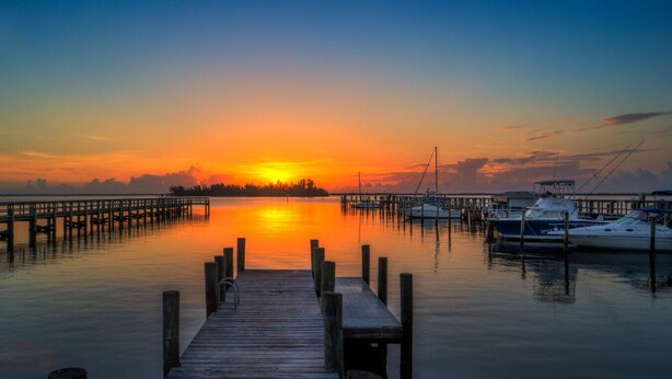 Een houten pier die zich uitstrekt over rustig water, met boten afgemeerd in een jachthaven, onder een zonsondergang.
