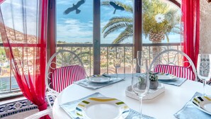 Un coin repas avec une table dressée pour deux personnes, des chaises dotées de coussins à rayures, une fenêtre offrant une vue sur des palmiers et le paysage urbain, et un ananas décoratif servant de centre de table.