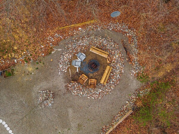 An aerial view of a circular outdoor seating area with a fire pit, surrounded by a gravel path and leaf-covered ground.
