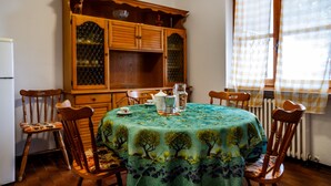 Un comedor con una mesa redonda cubierta con un mantel verde, sillas de madera, un mueble de madera con puertas de cristal y una ventana con cortinas a cuadros.