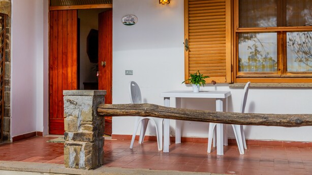 Una puerta de madera, un pilar de piedra, una mesa blanca con sillas y una ventana con contraventanas.