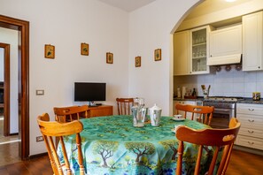 A dining area with a table covered by a floral tablecloth, surrounded by wooden chairs, a television, and kitchen cabinets.