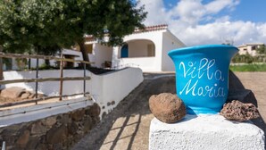A blue flower pot with 'Villa Maria' written on it, placed on a white concrete block with two rocks beside it, in front of a white building with a tree in the background. - Holiday Home 'Villa Maria' with Private Terrace and Private Garden (Orosei)