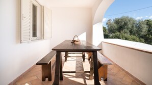 A balcony with a wooden table and benches, a window with shutters, and a view of trees. - Holiday Home 'Villa Maria' with Private Terrace and Private Garden (Orosei)