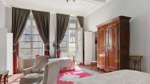 A hotel room with a large bed, a sitting area with a chair and a table, a wardrobe, and a view through the window. - Apartment 'La Suite Barbes' with Shared Garden, Balcony and Wi-Fi (Carcassonne)