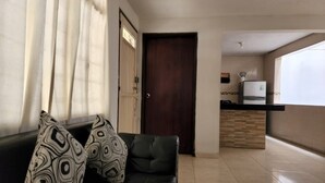 Una sala de estar con un sofá verde oscuro, cojines estampados, una puerta de madera y una zona de cocina con un refrigerador y una encimera.
