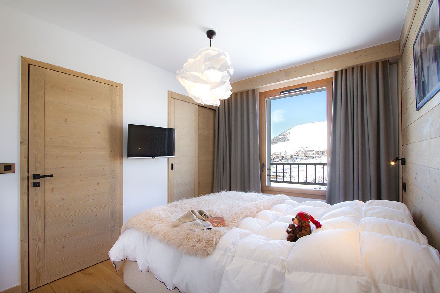 Une chambre avec un grand lit, un téléviseur fixé au mur, une fenêtre avec des rideaux et une vue sur un paysage enneigé.