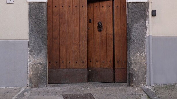 A wooden door with a metal handle, set within a white and grey wall with a decorative arch above. - De Vivo Realty - Fortuna House (Sant'Agnello)
