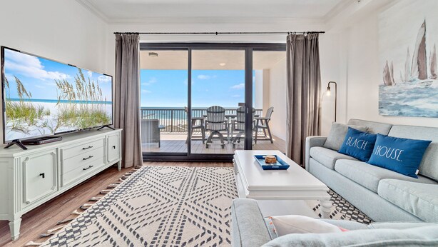 A living room with a large flat-screen TV, a sofa with 'Beach House' pillows, a patterned rug, and a balcony with a view of the ocean. - Grand Pointe 613 | Beachfront | Resort Amenities | MyBeach Getaways (Orange Beach)